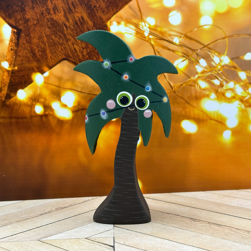 Decorative palm tree with googly eyes and colorful lights on a wooden surface with blurred lights in the background.