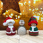 Three decorative figures with festive hats on a wooden surface with string lights in the background.