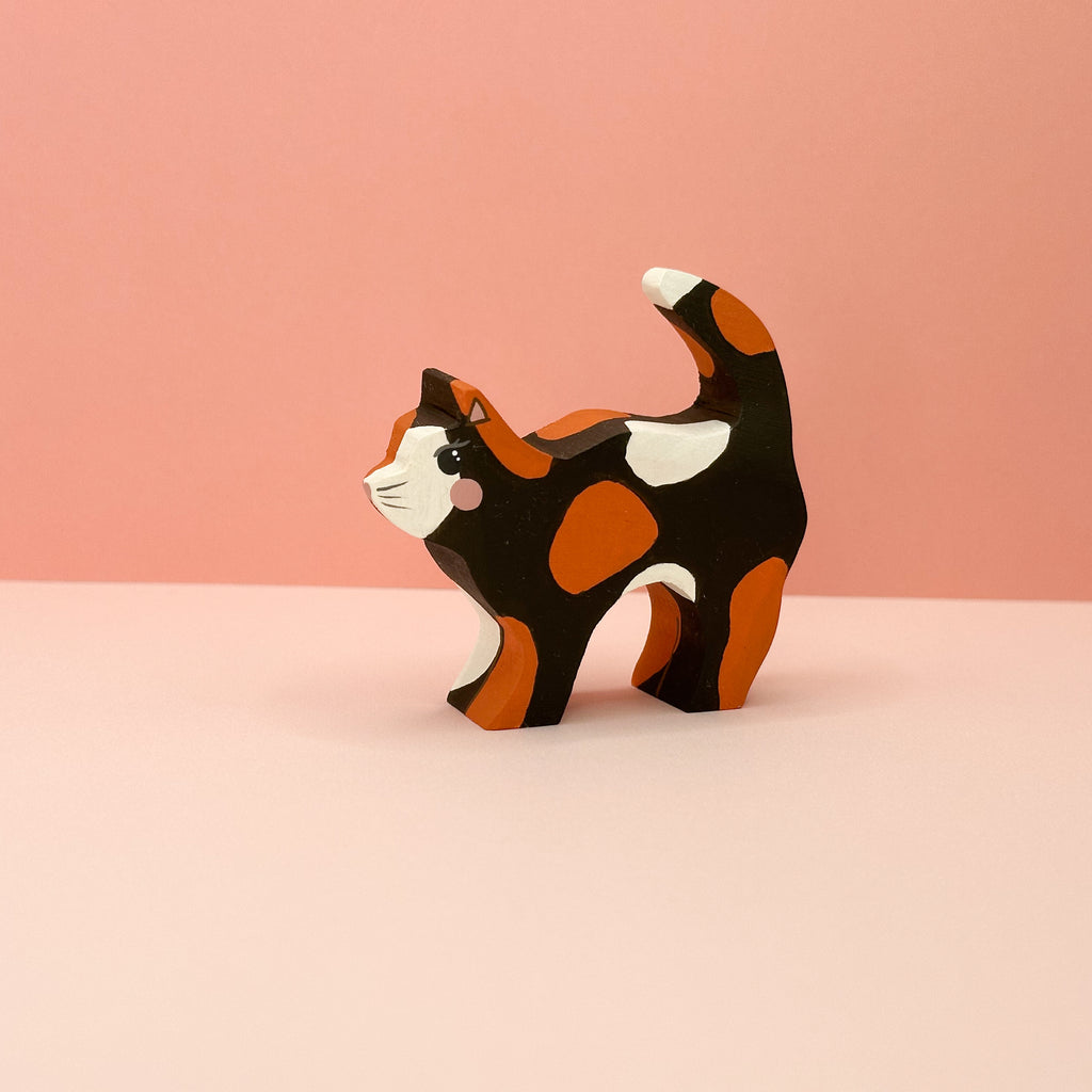 Wooden cat figurine with black, orange, and white pattern on a pink background